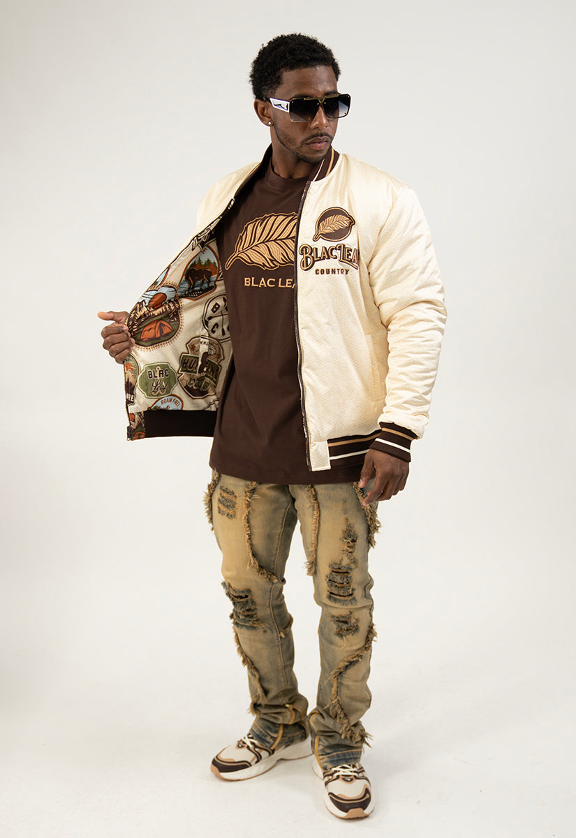 Man wearing a varsity jacket, brown shirt with khaki Leaf Embroidered, and ripped jeans on a white background
