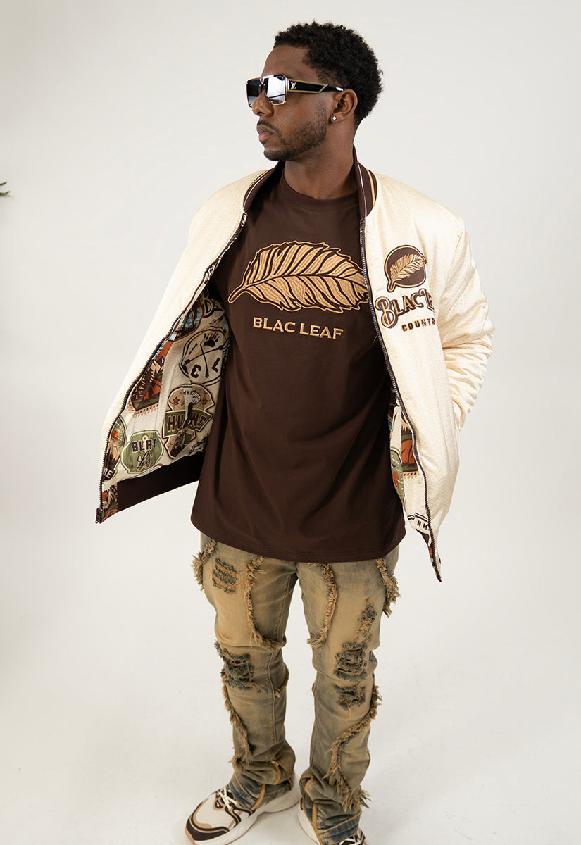 Man wearing a varsity jacket, brown shirt with khaki Leaf Embroidered, and ripped jeans on a white background